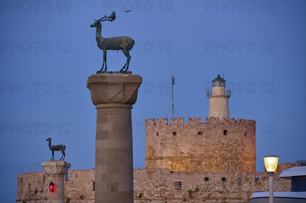 Bronze deer on pedestal in the foreground with old fortress walls and lighthouse in the background,