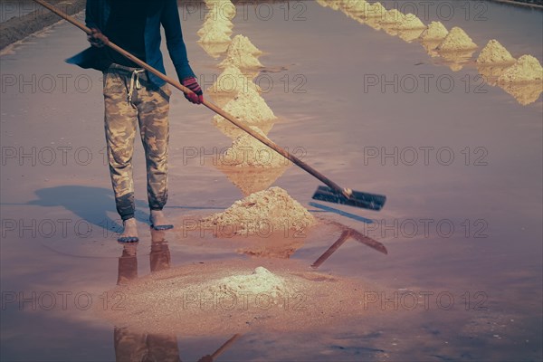 Low section of a khmer worker manually gathering salt into rows and heaps using a long wooden tool