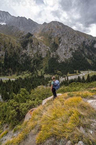 A female hiker stands on a grassy slope against an impressive mountain backdrop under a cloudy sky,