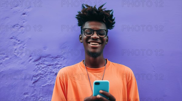 Black young positive man with dreadlocks is smiling and standing in front of the building wall.
