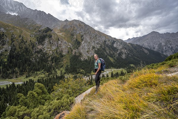 A female hiker stands on a grassy slope against an impressive mountain backdrop under a cloudy sky,