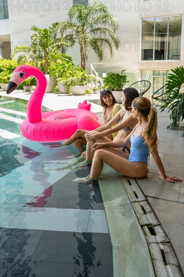 Three women are sitting and chatting on a poolside with a pink flamingo float. They are smiling and