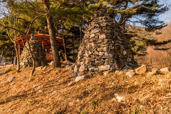 Two stone pyramids on small hill under grove of evergreen trees and wooden gazebo in background