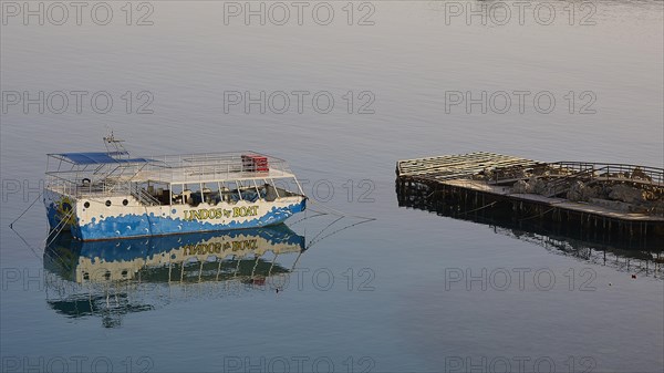 An excursion boat is reflected in the calm waters next to a wooden jetty, Lindos, Rhodes,