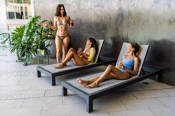 Two women are lounging in a courtyard, while the third is standing beside them. They are sipping on