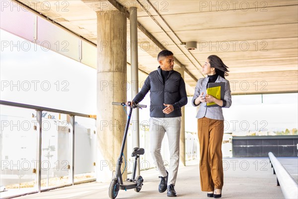 Woman in formal suit talking to man holding a scooter. Girl in formal suit talking to man holding