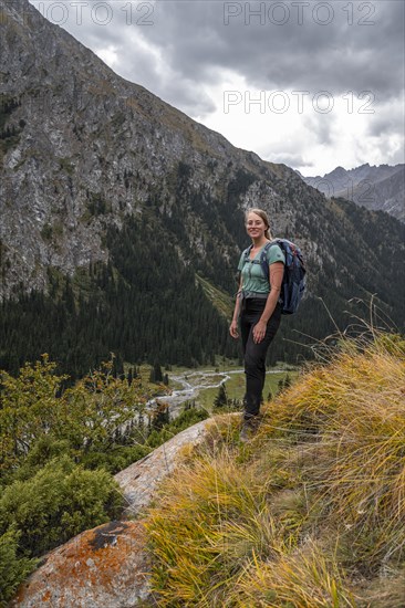 A female hiker stands on a grassy slope against an impressive mountain backdrop under a cloudy sky,