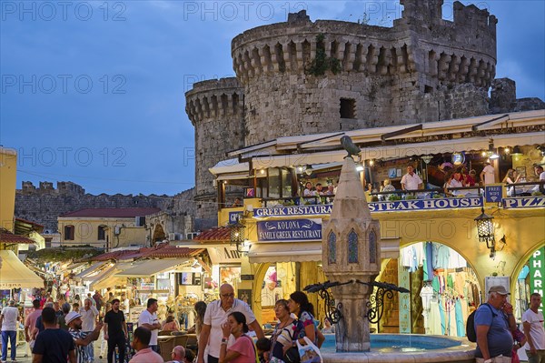 Hippocrates Square, St Paul's Gate, visitors walk between restaurants and shops by an old castle,