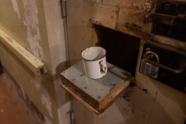 Rusted metal cup stands on the food hatch of a prison cell, former headquarters and prison of the