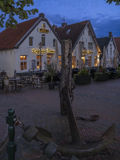 Ship's anchor on a wooden pile in front of restaurants at Greetsiel harbour at night, Greetsiel,