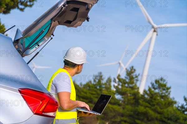 Male mature technician wearing reflective clothes and hardhat standing next to the car using laptop