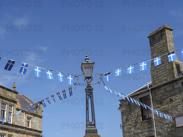 Street scene with blue and white flags hanging from a lamppost between old buildings, calm sea with