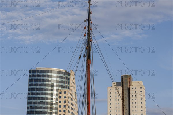 Sail mast between two modern buildings in front of a blue sky, skyline of a modern city on a river