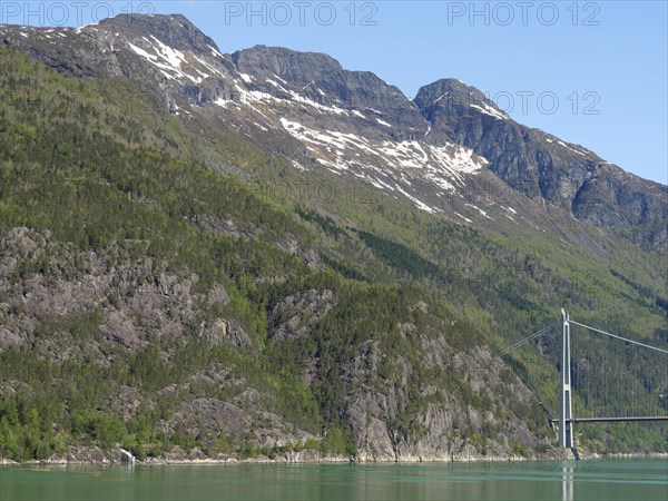 High mountains with remnants of snow on the peaks, green forested slopes and a large bridge under a