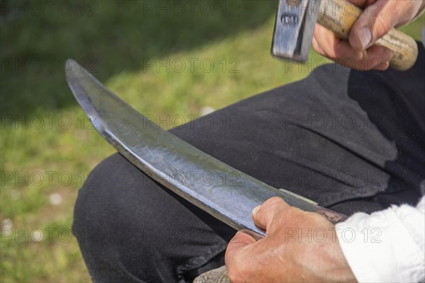 Man peening a scythe blade using a jig and a hammer to sharpen it