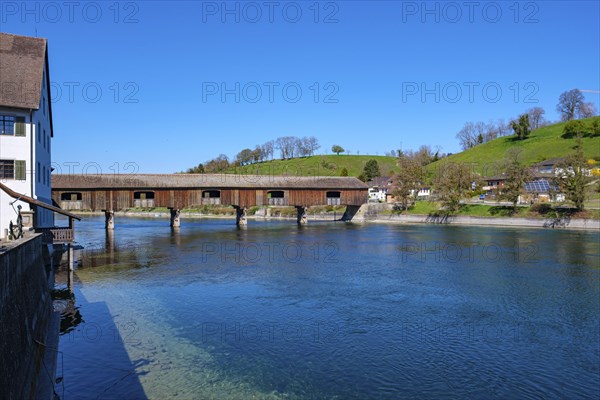 Wooden bridge over a river with old buildings in the background and a green meadow under a bright