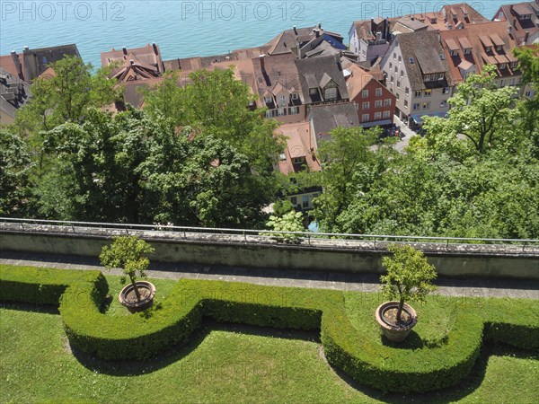 View of a picturesque garden with trees and trimmed hedges, in the background roofs and a lake, old