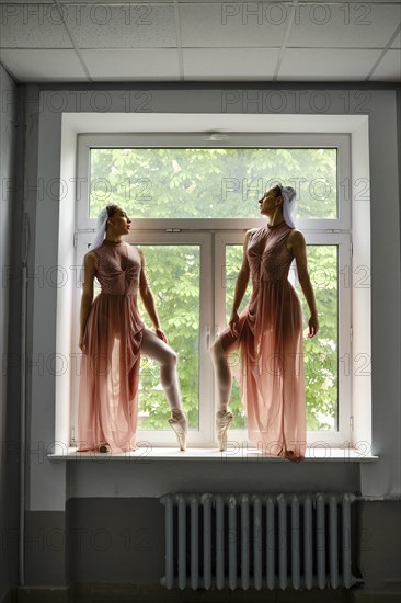 Two graceful ballerinas in transparent long dresses stand on the windowsill in the corridor