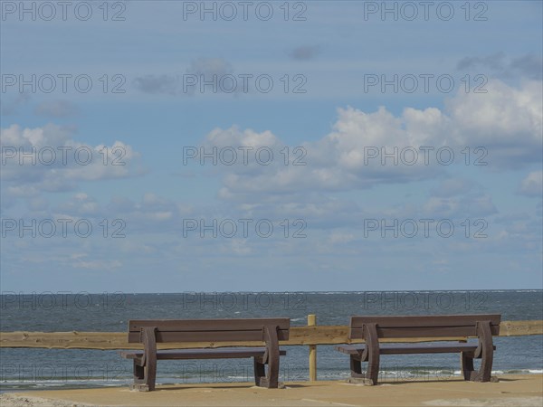 Two wooden benches overlooking the calm and wide sea under a cloudy sky, beach and dunes with grass