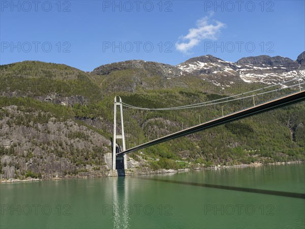 Massive bridge over a green lake, surrounded by forested and rocky mountains under a clear blue