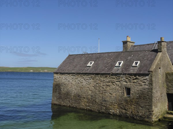 Small stone house on the shore of clear blue water with sweeping views of the coastline and blue