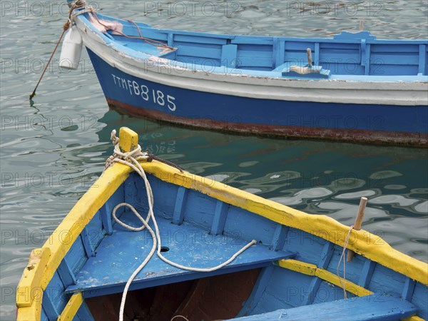 Two small boats in different colours floating in the harbour water, connected with ropes, many