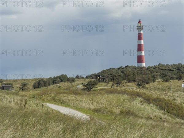 A red and white striped lighthouse in a grassy and wooded dune landscape under a cloudy sky, red