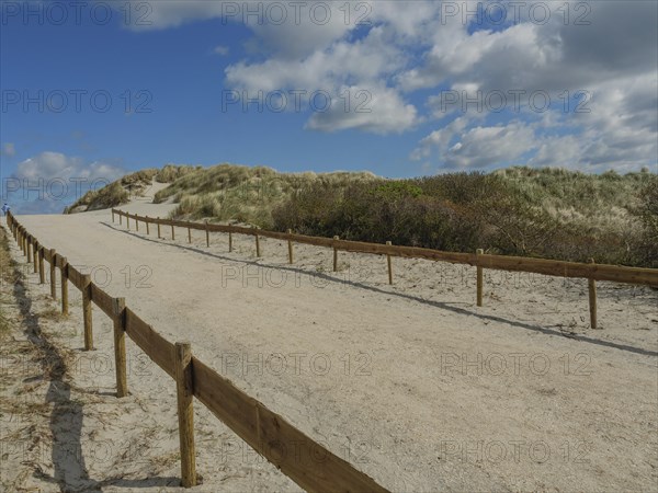 A sandy path lined with a wooden fence, sandy dunes and a sky with clouds, dunes on an island with