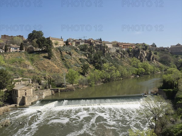A city view on a hill with buildings, a river with a waterfall and green areas under a clear sky,
