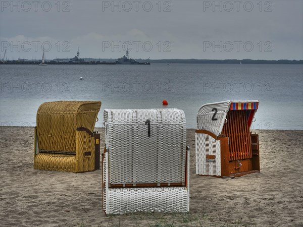 Three beach chairs on a sandy beach in front of the calm sea, ships on the horizon under a cloudy