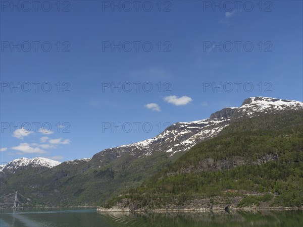 A clear day with snow-capped mountains under a blue sky with scattered clouds, bridge in a fjord