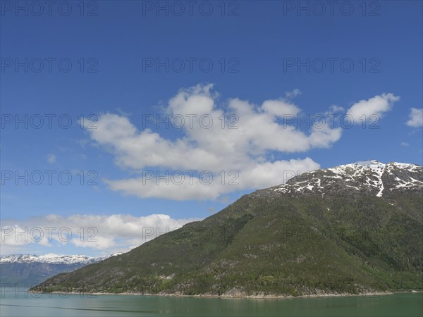 A mountain under a clear blue sky with some clouds, surrounded by green forests and a lake,