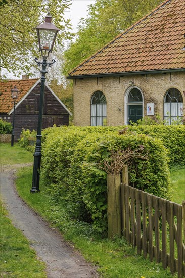 A house with a tiled roof and lamppost along a path, surrounded by green hedges, old houses with