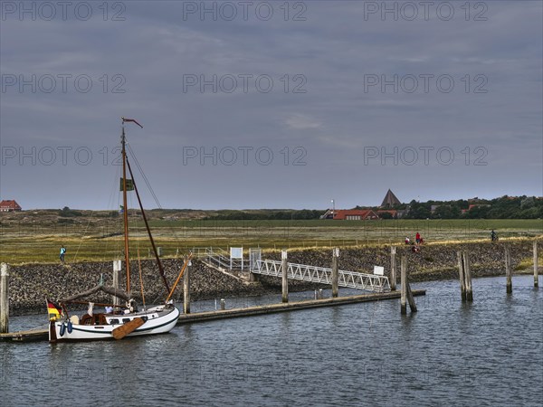 A sailing boat is moored at a pier, houses and people can be seen in the background, all under a