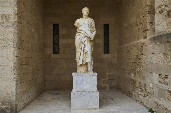 Ancient statue of a man in a long robe on a pedestal in front of a stone wall, torso from the 2nd