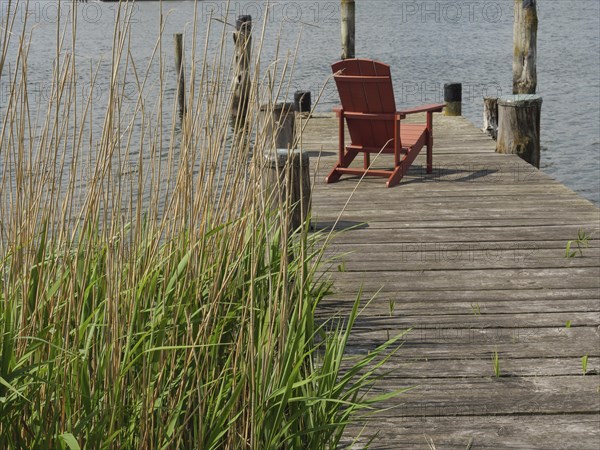 A quiet wooden jetty on the lakeshore with a single red seat and green reeds in the foreground,