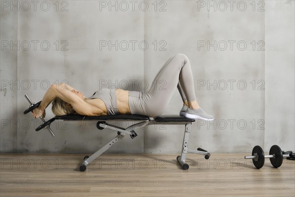 A muscular woman lies on a bench in the gym and pushes a dumbbell behind her head with both hands