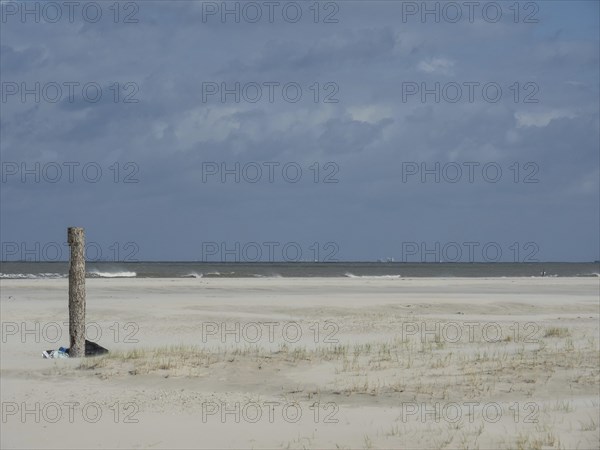 Lonely beach with dunes in front of a grey, cloudy sky. The sea is calm and spacious, waves on a