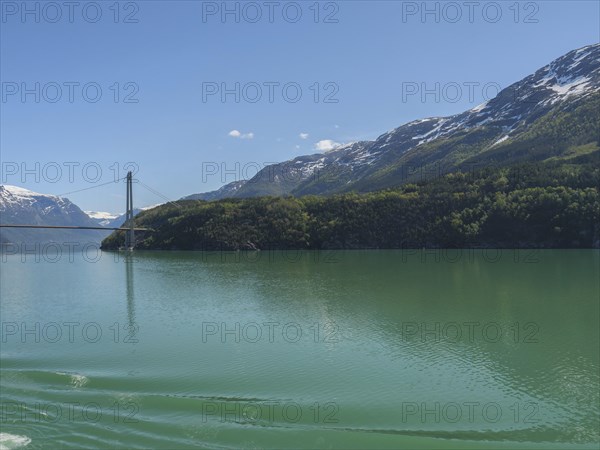 Turquoise lake surrounded by snow-capped mountains, a bridge spans in the background under a blue