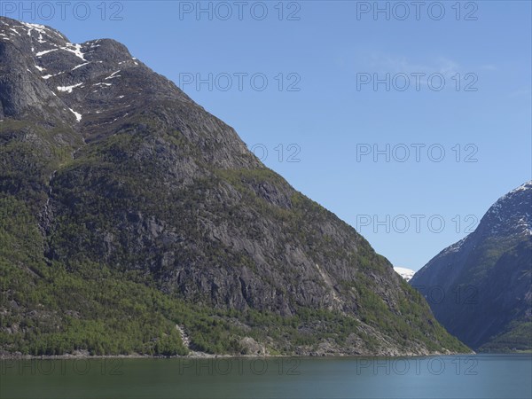 Steep rock faces covered with a forest reach down to a clear water and snow-capped mountain peaks