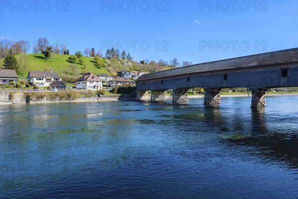 Massive wooden bridge over a clear river, surrounded by houses and green hills, under a blue sky,