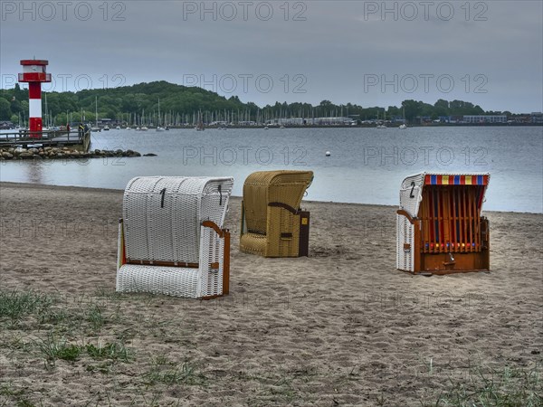 Several beach chairs standing on a cloudy beach, behind them a lighthouse and calm sea under a