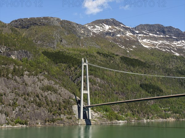 Bridge spanning forested mountains and a green lake, under a clear blue sky, bridge in a fjord with