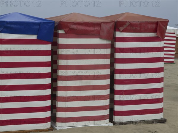 Three colourful beach huts with striped pattern stand on the sandy beach, beach chairs and beach