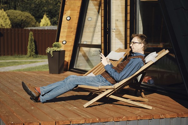 Middle aged man using smartphone while resting on terrace of wooden cabin in a sunny day
