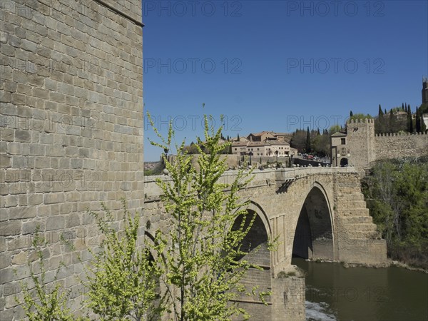 Historic stone bridge crossing a river, surrounded by green vegetation and clear blue sky, historic