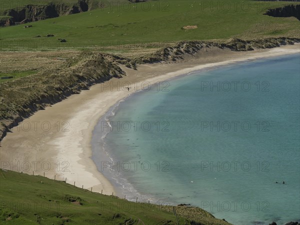 A quiet sandy beach surrounded by rocky cliffs and gentle green slopes, with turquoise clear water