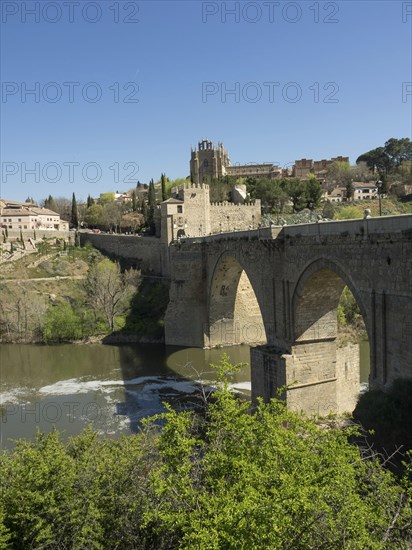 Stone bridge with arches over a river, surrounded by a cityscape and vegetation, sunny weather,