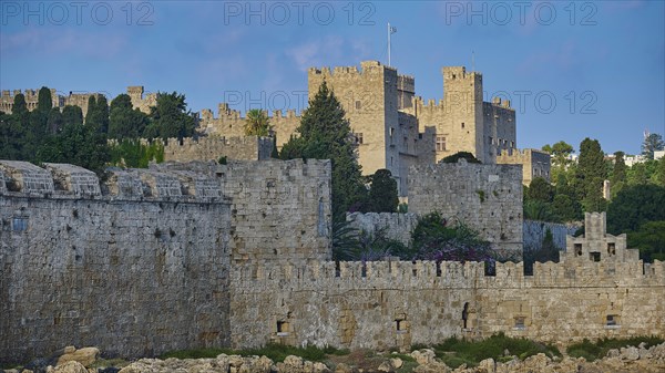 Ancient fortress walls with battlements, surrounded by lush greenery under a partly cloudy sky,