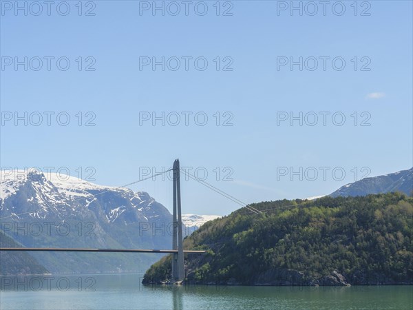 Landscape with high bridge, snow-capped mountains and green wooded hills under a clear blue sky,